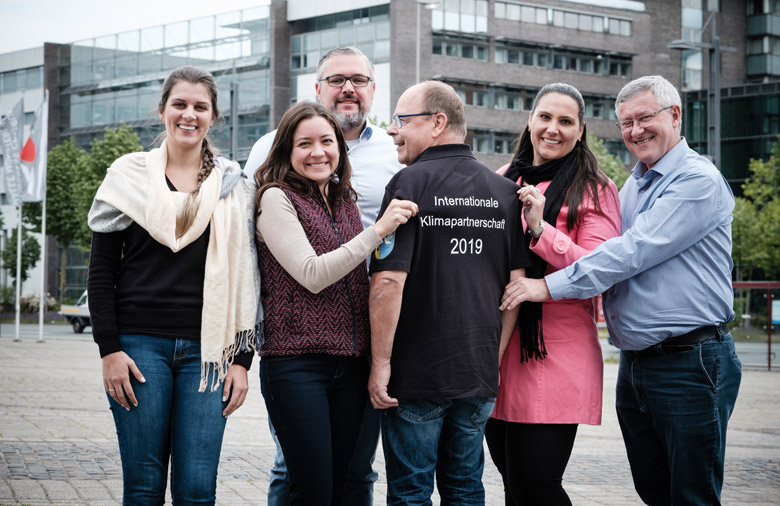 Sechs lachende Personen sind zu sehen; im Hintergrund ein modernes Gebäude. Ein Mann steht mit dem Rücken zum Betrachter. Hinten auf seinem Shirt steht: Internationale Klimapartnerschaft 2019.